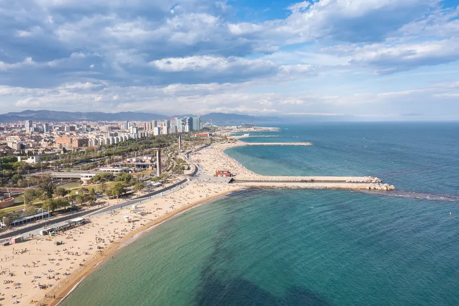 A wide sandy city beach and breakwaters seen from the air, turquoise water running out to the open sea under white clouds