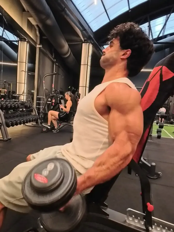 Gio doing incline dumbbell curls on a bench in a bright gym.