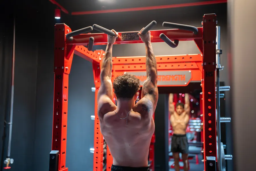 Gio from behind, hanging from the pull-up handles of a power rack in front of a mirror.