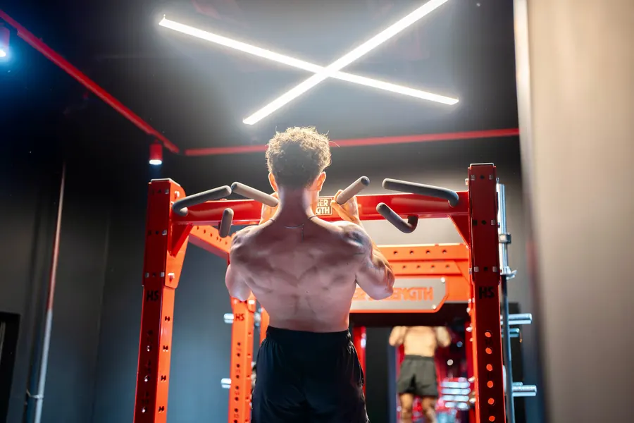 Gio from behind gripping a pull-up bar under a cross of white ceiling light, back muscles lit from above