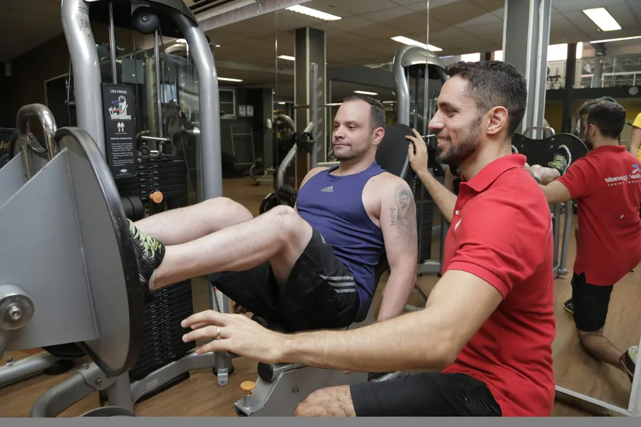A trainer in a red polo kneels beside a man on a leg press, guiding the movement with his hand